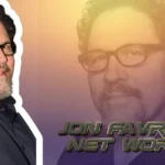 Jon Favreau smiling in black suit and glasses with 'Jon Favreau Net Worth' text overlay for celebrity wealth article