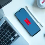 gadgetfreeks.com: smartphone showing Netflix logo on desk with laptop, stylus, and earbuds in a minimalist tech flat lay