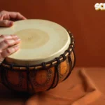 Hands playing scimitar drum with ornate decorative patterns on terracotta background showing proper finger technique