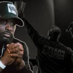 Philip Anthony Mitchell net worth - 2819 Church pastor preaching with hands clasped in prayer wearing baseball cap and hoodie