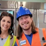 Handyman Hal (Shawn Goins) in blue hard hat and safety vest smiling with colleague in kitchen