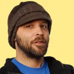 Brian Redban wearing a beanie and jacket against a yellow background, representing redban net worth and his comedy career.