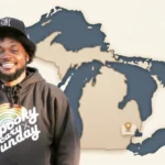 CoryxKenshin smiling in a Spooky Cary Sunday hoodie standing in front of a Michigan state map with a location pin on Farmington Hills
