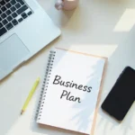 Minimalist home office workspace with laptop, business plan notebook, smartphone, and coffee showing low investment business startup setup